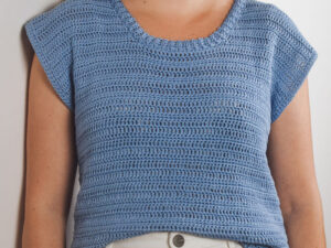 Une personne portant un haut bleu clair à manches courtes et à encolure ronde, associé à un pantalon blanc à taille haute - un look élégant parfait pour toute personne cherchant à s'inspirer d'un tuto de t-shirt au crochet. L'arrière-plan est uni et de couleur claire.