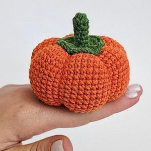 Une petite citrouille orange, réalisée au crochet et dotée d'une tige verte, est tenue entre les doigts d'une personne, sur un fond blanc uni.
