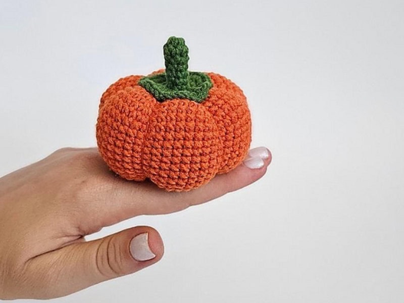 Une main aux ongles vernis de blanc tient une petite « citrouille au crochet » — une citrouille orange tricotée au crochet, dotée d'une tige verte — sur un fond blanc uni.