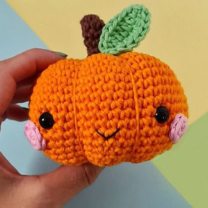 Une main tient une citrouille au crochet, avec des yeux en boutons noirs, des joues roses, un petit sourire, une tige marron et une feuille verte, sur un fond aux couleurs pastel.