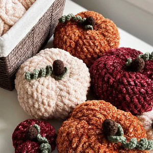Une collection de citrouilles crochetées dans des tons crème, orange et rouge, avec des tiges vertes et marron, disposées à côté d'un panier tressé sur une surface blanche.