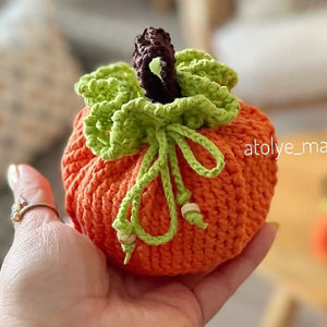 Une main tient une citrouille orange réalisée au crochet, dotée d'une tige marron et de feuilles vertes, avec un cordon vert noué autour du haut à des fins décoratives. L'arrière-plan est légèrement flou.