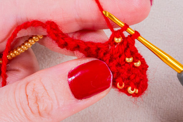 Gros plan sur une main aux ongles vernis en rouge, en train de crocheter avec du fil rouge et des perles dorées à l'aide d'un crochet doré, illustrant comment crocheter avec des perles avec élégance.