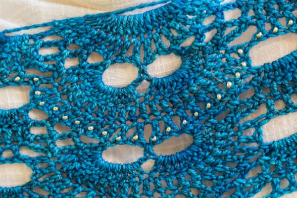 Gros plan sur une dentelle au crochet bleue aux motifs complexes, circulaires et ajourés, posée sur un fond blanc : une source d'inspiration idéale pour celles et ceux qui aiment crocheter avec des perles.