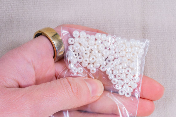 Une main ornée d'une bague en or tient un petit sachet en plastique transparent rempli de perles blanches rondes, idéales pour le crochet avec des perles, sur un fond beige.