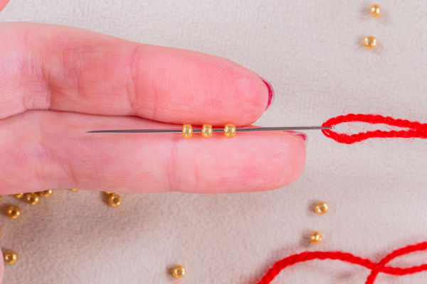 Gros plan sur des doigts tenant une aiguille enfilée d'un fil rouge et de trois petites perles dorées, illustrant comment crocheter avec des perles, avec d'autres perles dorées éparpillées sur une surface claire en arrière-plan.