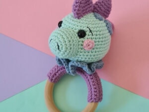 Un charmant hochet au crochet en forme de licorne, avec une tête vert menthe, une crinière et des oreilles violettes, des yeux noirs et un blush rose, le tout monté sur un anneau en bois, le tout sur fond aux couleurs pastel.