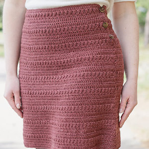 Une personne vêtue d'un haut blanc et d'une jupe en crochet rose poudré, texturée et à taille haute, ornée de boutons sur le côté, se tenant debout à l'extérieur, les bras le long du corps.