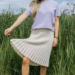 Une femme se tient debout dans un champ d'herbes hautes ; elle porte un t-shirt lavande et une jupe plissée de couleur crème. On ne voit pas son visage, et elle tient sa jupe d'une main. Le ciel est partiellement nuageux.