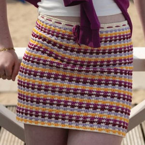 Une personne portant une mini-jupe au crochet à rayures horizontales violettes, jaunes, crème et blanches, debout à l'extérieur, avec une partie d'une balustrade blanche visible.