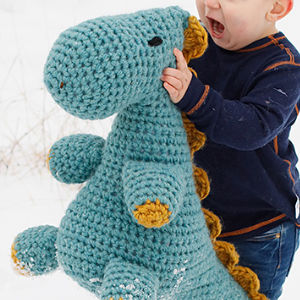 Un petit enfant vêtu d'un t-shirt bleu marine serre contre lui une grande peluche de dinosaure bleue tricotée au crochet, ornée de touches de jaune, à l'extérieur, dans la neige.