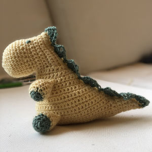 Un jouet dinosaure crocheté avec un corps jaune et des accents verts, notamment des pointes le long de son dos, de ses pattes et de sa queue, est posé sur une surface claire avec un arrière-plan neutre.