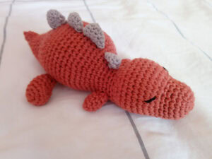 Un jouet dinosaure orange fait main au crochet, inspiré d'un modèle gratuit de dinosaure au crochet, avec les yeux fermés et trois petites pointes grises le long du dos, couché sur une couverture blanche avec de fines lignes grises.