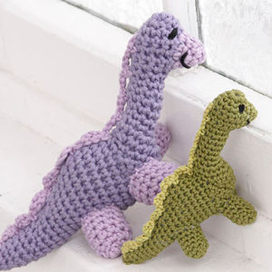 Deux jouets en crochet représentant des dinosaures, l'un violet et l'autre vert, sont placés côte à côte sur un rebord de fenêtre blanc, tous deux tournés vers la fenêtre.