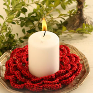 Une bougie cylindrique blanche repose sur une assiette en argent, entourée d'un napperon rouge au crochet. Des branches aux feuilles vertes se détachent en arrière-plan.