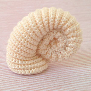 Un objet au crochet, de couleur crème, aux contours souples, en forme de coquille d'ammonite enroulée, posé sur une surface texturée de couleur rose pâle.