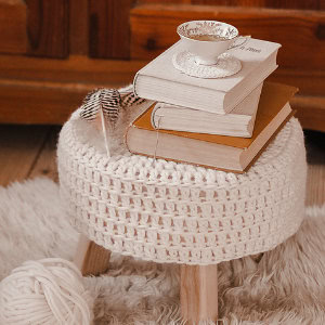 Un petit tabouret rond recouvert d'un napperon blanc au crochet supporte une pile de livres, une tasse à thé posée sur une soucoupe et deux plumes rayées. Le tabouret est posé sur un tapis moelleux, à côté d'une pelote de laine blanche.