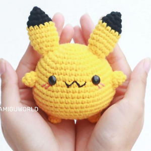 Une paire de mains tient une petite peluche jaune, ronde et crochetée, avec des oreilles noires, des joues rougissantes et un visage mignon ressemblant à celui de Pikachu.