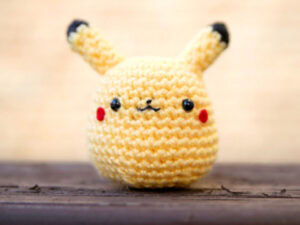 Une petite poupée pikachu au crochet avec du fil jaune, des oreilles noires, des joues rouges et un visage simple et souriant est posée sur une surface en bois sur un fond flou.