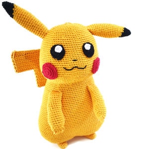 Poupée Pikachu au crochet, faite à la main, avec un corps jaune, des oreilles noires, des joues rouges et des yeux noirs, se tenant debout sur un fond blanc uni.