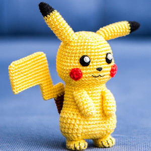 Une figurine crochetée de Pikachu jaune avec des oreilles noires, des joues rouges, de grands yeux et une queue en zigzag, se tenant debout sur une surface bleue avec un fond bleu.