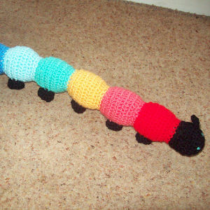 Une chenille en crochet avec une tête noire et des segments de corps colorés en rouge, rose, jaune, vert, bleu et bleu clair, posée sur un tapis beige.