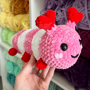 Une main tient une chenille en peluche, rose et blanche, crochetée, avec un visage souriant, des pieds rouges et des antennes rouges en forme de cœur. Des écheveaux de laine de différentes couleurs sont visibles à l'arrière-plan.
