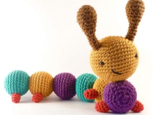Une chenille au crochet avec une tête jaune, des antennes brunes, des segments de corps bleus et violets, et de petites pattes rouges sourit sur un fond blanc - parfait pour suivre un tuto chenille au crochet gratuit.
