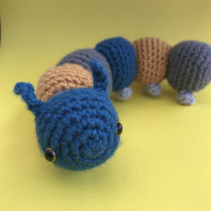 Une chenille crochetée avec des segments bleus, bruns et gris et de petits yeux noirs est posée sur un fond jaune.
