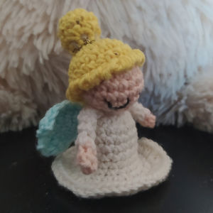 Une petite figurine d'ange au crochet, coiffée d'un chapeau jaune, dotée d'ailes bleues et vêtue d'une robe blanche, assise sur une surface sombre avec un fond crème duveteux.