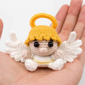Un petit ange au crochet, avec une auréole jaune et des ailes blanches, est posé dans la paume d'une main.