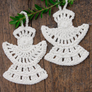 Deux décorations en forme d'anges, crochetées en blanc et ornées de motifs ajourés, reposent sur une surface en bois. Une petite branche verte feuillue est posée au-dessus des décorations.