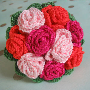 Gros plan sur un bouquet composé de fleurs au crochet dans des tons de rose, de rouge et de fuchsia, agrémenté de feuilles vertes au crochet, disposées en une composition ronde sur un fond clair.