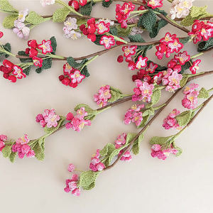 Des fleurs de cerisier au crochet, roses, rouges et blanches, accompagnées de feuilles vertes, disposées sur des tiges marron, sur un fond clair.
