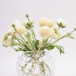 Un vase en verre transparent et texturé contient un bouquet de fleurs artificielles vert clair et blanches, composé notamment de pompons jaunes ronds tricotés au crochet et de boutons de renoncule, sur un fond blanc uni.