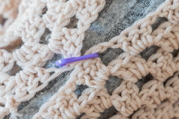 Gros plan sur un motif de couture au crochet de couleur beige, orné d'un marqueur de mailles en plastique violet, posé sur un fond en tissu gris et blanc.