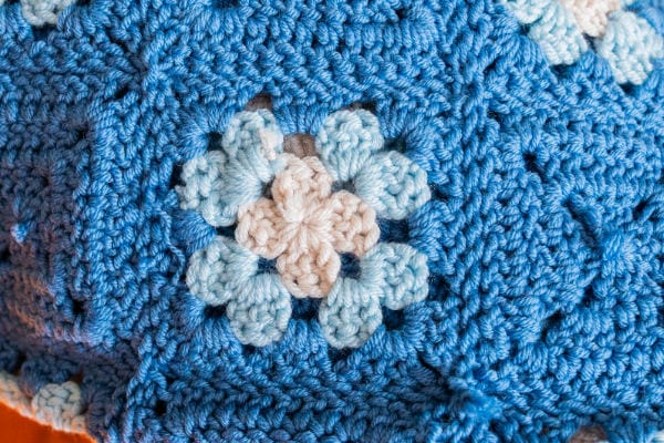 Gros plan sur une couverture « granny square » réalisée au crochet, ornée d'un motif floral en fil blanc, bleu clair et bleu moyen. Les mailles et la texture du travail au crochet sont clairement visibles.