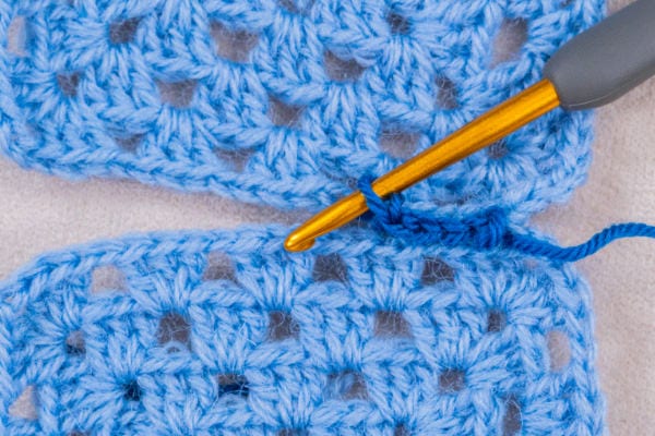 Un crochet doré assemble deux morceaux de tissu au crochet bleu clair avec du fil assorti, permettant d'observer de près le processus de réalisation de la « couture au crochet ».