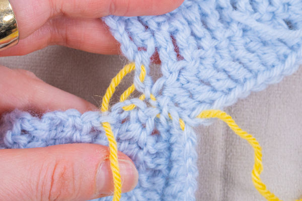 Gros plan sur des mains en train de coudre ensemble deux morceaux de tricot bleu clair à l'aide d'un fil jaune vif et d'une aiguille, illustrant une technique de couture ou d'assemblage dite « couture au crochet ».