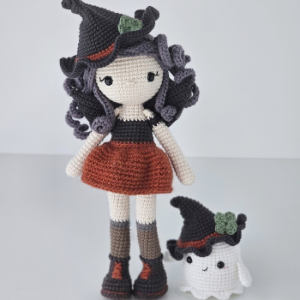 Une poupée au crochet aux cheveux bouclés, coiffée d'un chapeau de sorcière, vêtue d'un haut noir, d'une jupe orange et de bottes, se tient à côté d'un petit fantôme au crochet portant un chapeau de sorcière assorti, rehaussé d'une touche de vert.