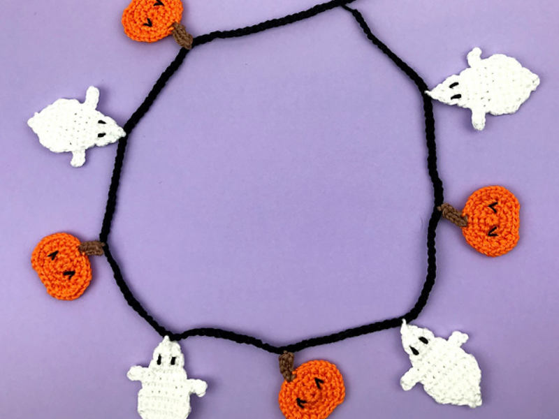 Une guirlande d'Halloween réalisée au crochet : d'adorables fantômes blancs et des citrouilles orange, tous crochetés, disposés en cercle sur un fond violet.