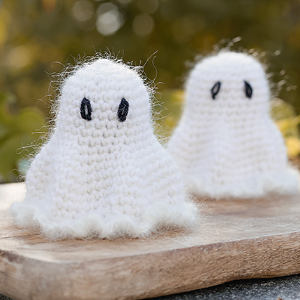 Deux petits fantômes blancs, tricotés au crochet et aux yeux noirs, sont posés sur une surface en bois à l'extérieur, avec un arrière-plan naturel flou.