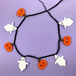Guirlande au crochet composée d'alternances de citrouilles orange et de fantômes blancs, suspendue à un fil noir, présentée sur un fond violet.