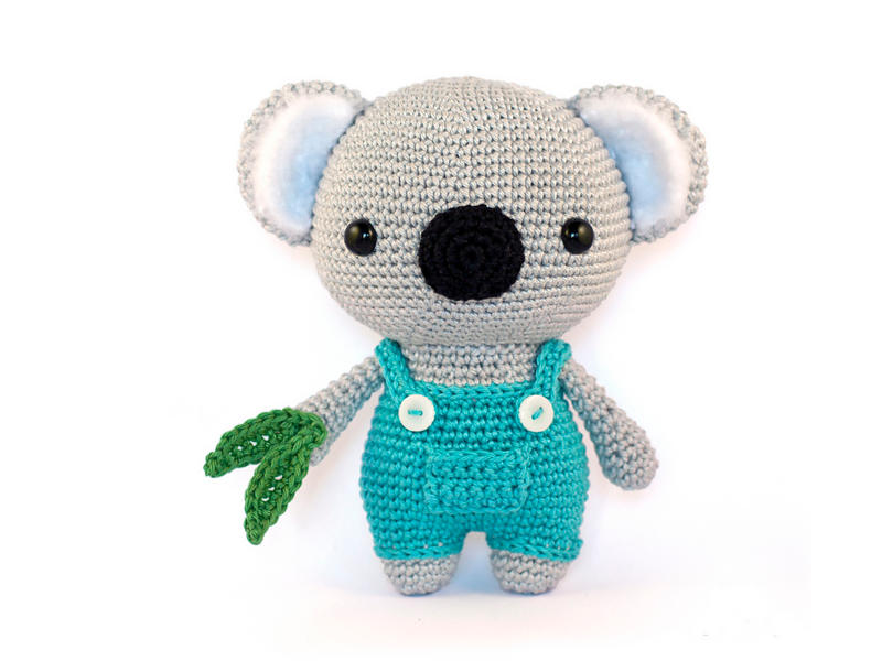 Une adorable peluche de koala au crochet porte une salopette bleu sarcelle à boutons blancs et tient dans ses mains une petite feuille verte également crochetée, le tout sur fond blanc.
