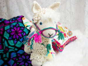 Un lama au crochet, orné de pompons colorés sur son harnais, est posé sur une surface blanche, à côté de coussins aux motifs éclatants et d'une couverture au crochet.