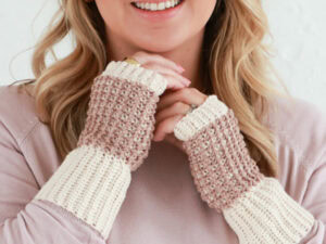 Une femme souriante porte d'élégantes mitaines au crochet — des mitaines sans doigts de couleur crème et marron clair — assorties à un haut rose pâle.