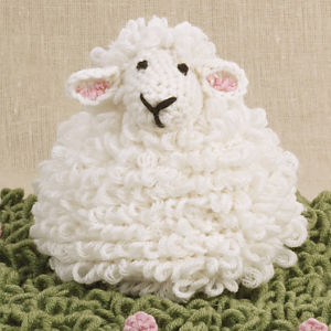 Un mouton en laine blanche duveteuse, crocheté à la main, aux yeux noirs et au sourire, est posé sur un tissu vert texturé parsemé de petites bosses roses, sur fond beige.
