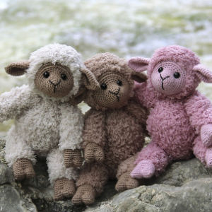Trois agneaux en peluche — un blanc, un marron et un rose — sont assis tout près les uns des autres sur un rocher, avec un arrière-plan extérieur flou.
