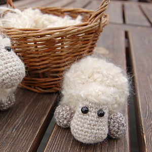 Deux petits moutons au crochet, à la laine duveteuse et aux yeux en boutons noirs, sont posés sur une table en bois, avec en arrière-plan un panier en osier rempli de laine du même genre.