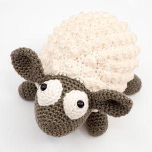 Une peluche de mouton au crochet, avec un corps texturé de couleur crème et une tête, des oreilles, des pattes et une queue gris foncé, dotée de grands yeux blancs aux pupilles noires, sur un fond blanc uni.